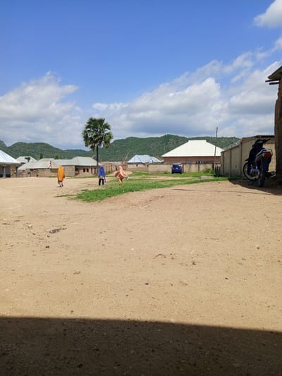 A low-angle, eye-level outdoor shot captures a sunny day in Barakin Babele, Nigeria. The scene is dominated by a dusty, light brown earth foreground, with a dark shadow cast from the bottom left corner. In the mid-ground, three individuals are walking across a patch of green grass. Two are women wearing long, flowing garments. The woman on the left wears a bright orange hijab and skirt, while the woman in the middle wears a blue hijab and dark clothing. The woman on the right is further away and wears a light brown or pinkish outfit, her right arm raised. Behind them, a cluster of buildings with corrugated metal roofs are visible, some with light-colored walls. A tall palm tree stands prominently behind the middle woman. In the background, green, rolling hills under a bright blue sky with scattered white clouds form the backdrop. To the right of the frame, a concrete wall with barbed wire runs along the property line, and a dark blue motorcycle is parked against a brick wall. Further down, a dark blue car is parked on a patch of grass in front of a building with a white, angled roof. The overall impression is a rural or semi-rural setting on a clear, pleasant day, likely in the late morning or early afternoon given the bright sunlight and the shadow cast.