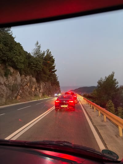 The image shows a view from inside a vehicle traveling on a two-lane road in Košljun, Montenegro. The road is bordered by a steep, rocky embankment on the left, covered in dense green vegetation. On the right, a yellow metal guardrail separates the road from a downward slope with trees. Several cars are visible ahead, appearing to be in a traffic queue. The taillights of these vehicles are illuminated, casting a red glow on the wet road surface. The sky is a pale, hazy gray, suggesting it is either early morning or late evening. In the distance, a body of water and hills are faintly visible, characteristic of the coastal landscape of Montenegro. The foreground shows the dashboard and windshield wipers of the vehicle.