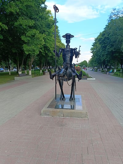 The image shows a metallic sculpture of a man on a horse in a park-like setting. The sculpture is in the foreground, positioned on a paved walkway. The man is wearing a hat and holding a spear. The horse appears to be made of similar metallic materials. Behind the sculpture, the walkway continues, lined with trees and benches. Several people are visible in the background, some sitting on benches and others walking. Cars are parked along the road in the distance. The sky is blue with scattered clouds. A sign with text is visible at the base of the sculpture. Based on the inscription on the sculpture's base, the location is identified as a monument to G.Y. Kuprianov in the city of Gubkin, Russia.