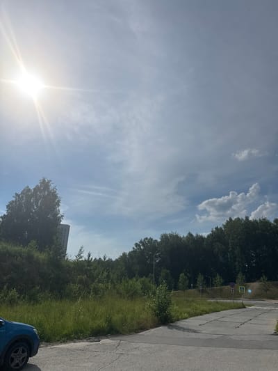 The image depicts a sunny outdoor scene with a bright sun in the upper left corner, casting strong rays across a pale blue sky with scattered clouds. In the foreground, a portion of a blue car is visible on the left. A paved road or parking area occupies the bottom right of the frame, with patches of grass and vegetation growing on either side of it. A dense line of trees forms a backdrop across the middle ground, with a tall, light-colored building partially visible behind a large tree on the left. Further back, several road signs are present, including a pedestrian crossing sign and a parking sign. The overall environment appears to be a suburban or semi-urban area on a clear day. The specific location is identified as Koltsovo, Russia.