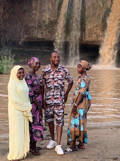 Four people stand on a sandy bank in front of a waterfall. The person closest to the camera wears a light yellow head covering and long dress. A woman in a black and pink patterned dress and head wrap stands next to her. A man stands next to her, wearing a patterned shirt and shorts. Furthest from the camera, a woman in a brown and turquoise patterned dress and head wrap stands. The waterfall is in the background. This scene is in the city of Kukum, Nigeria.