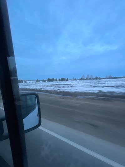 The image, likely taken from inside a vehicle, shows a flooded landscape alongside a road. The road itself is partially submerged, with water reaching up to what appears to be a line of trees or vegetation in the distance. A rearview mirror is visible within the vehicle. The sky is overcast with a blue hue. The reflection visible in the rearview mirror does not provide clear details. The location of the photo is in the city of Povarenki, Russia.