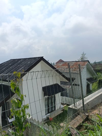 Two white houses with dark and red roofs stand side by side, partially obscured by a wire fence. The front house features dark-framed windows and a smaller roof over the entrance. A bit of green vegetation is visible in the foreground, growing along the fence. The sky above is filled with puffy white clouds, suggesting a partly sunny day. The setting appears to be a hillside or elevated area, with glimpses of more greenery in the background.