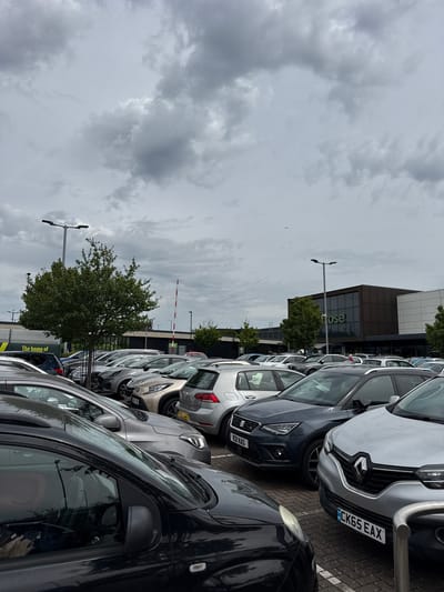 The image shows a car park adjacent to a large building, likely a retail store, in High Wycombe, United Kingdom. Several cars are parked closely together. The sky is overcast with grey clouds. In the background, a tree and a barrier are visible. The building is multi-story, with a glass facade bearing the word "Waitrose". Street lamps are visible throughout the car park.