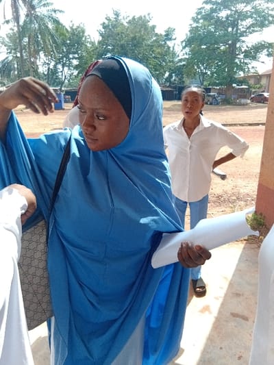 A woman in a blue hijab stands in the foreground, looking down and gesturing with her left hand. She carries a patterned handbag on her shoulder. Behind her, another woman in a white shirt and blue pants stands with a hand raised and a rolled-up paper or document in her other hand. They appear to be outdoors, possibly near a building with trees visible in the background.