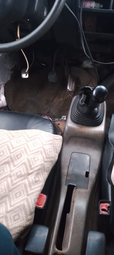 The media file shows the interior of a vehicle, likely a car. The scene focuses on the driver's side footwell, the gear shift console, and a portion of the passenger seat with a patterned cushion.
In the footwell, three pedals are visible: the clutch pedal on the left, the brake pedal in the middle, and the accelerator pedal on the right. Wires are also present, one of which is hanging down near the clutch pedal. The floor appears to be covered with a dark, possibly rubber, mat, and the overall condition of the floor and surrounding areas suggests wear and tear.
The gear shift console is prominent, with a black gear shift knob and a rubber gaiter. To the right of the gear shift, there's a storage compartment with a lid. The central console appears to be made of plastic and shows signs of use. On the passenger side, a portion of a dark seat is visible, covered partially by a light-colored cushion with a geometric diamond pattern. Red seatbelt buckles are also visible on either side of the console.
The lighting suggests it is daytime, but the interior of the car is somewhat dimly lit. No specific weather conditions are discernible. The context "Jibowu, Nigeria" indicates the geographical location. The image does not clearly depict any people or specific activity other than the interior of a stationary vehicle. There are no discernible signs or text in the image. The overall impression is of a used vehicle interior, possibly in need of cleaning or repair.