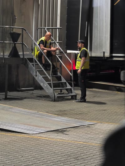 At a loading dock in Zeppelinheim, Germany, two individuals, both wearing high-visibility vests, are present. One person is seated on a metal staircase, looking at a mobile phone. The other individual stands nearby, facing the first person. Behind them, a large trailer with metallic siding is visible. The ground is paved with interlocking bricks, and a metal ramp with a patterned surface is in the foreground. The lighting suggests it is nighttime.