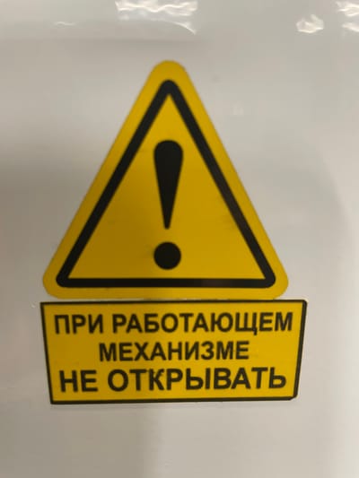 The image shows a yellow triangular warning sign with a black border and a black exclamation mark in the center. Below the sign, there is a yellow rectangular label with black text in Russian. The text reads: "ПРИ РАБОТАЮЩЕМ МЕХАНИЗМЕ НЕ ОТКРЫВАТЬ", which translates to "DO NOT OPEN WHILE THE MECHANISM IS RUNNING". The sign is affixed to a white surface, which appears to be a wall or a piece of equipment. The background is mostly white with some subtle shadows and reflections. There are no people or animals visible in the image. The image appears to be a close-up shot of the warning sign and label. There is no information in the image that can be used to determine its location in the city of Терволово, Russia.