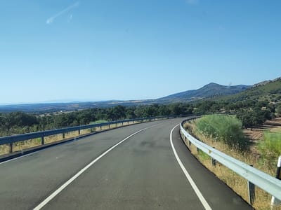 The image shows a view from a vehicle traveling on a paved, winding road through a rural, hilly landscape. The road has white lane markings and is bordered by metal guardrails and dry grass. In the background, there are rolling hills covered with trees and sparse vegetation, and a clear, light blue sky. The scene is brightly lit by sunlight, suggesting daytime. This appears to be a view of a road outside of Valencia de Alcántara, Spain, given the characteristic landscape of the region. There are no people or specific structures visible in the immediate foreground or on the road itself. The overall impression is one of a scenic drive through a dry, Mediterranean-like environment.