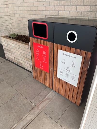 This media file captures a modern, two-compartment recycling and waste bin, likely situated in a public or commercial outdoor setting in Indooroopilly, Australia. The bin has a sleek design, featuring a black metal frame with two front panels adorned with horizontal wooden slats.

The upper compartment is for "Bottles & Cans," indicated by a circular opening with a white rim. A white instructional sign on the wooden panel lists the acceptable items with corresponding icons: plastic and glass bottles, aluminum and steel cans, used juice and milk containers (cartons), and clean paper and plastic cups. The sign emphasizes "Recyclable items ONLY."

The lower compartment is designated for "Landfill," marked by a rectangular opening with a red rim. A red sign provides guidance for this section, showing icons for food waste, soft plastic and plastic bags, coffee cups, food wrappers, and general waste. Both informational signs prominently display the branding "INDOOROOPILLY HOTELS."

The bin stands on a paved surface composed of different types of tiles: large, light grey square tiles on the right and smaller, lighter beige rectangular bricks on the left, separated by a metal strip embedded in the ground. Adjacent to the landfill compartment, a small planter box containing some sparse, dry-looking greenery is visible. The lighting is bright and even, indicating daytime conditions. No people are present in the image, and no specific event is taking place, presenting a static view of the waste disposal unit.