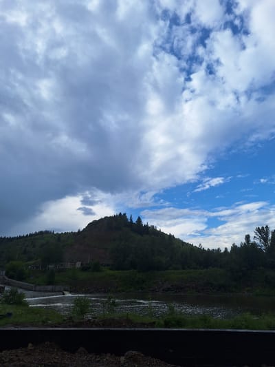 The image shows a landscape view of the river Sim near the city of Sim, Russia. A dam is visible on the left side of the image, with water flowing over it. A body of water stretches across the middle of the frame, reflecting the sky. In the background, a lush green hill is covered with trees. The sky is partly cloudy with patches of blue visible. The foreground shows rough terrain with some rocks and debris.