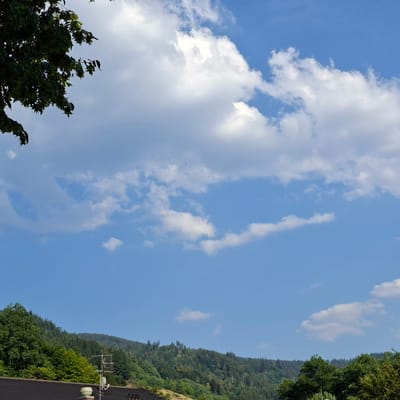 This is a medium shot of a bright, partly cloudy sky over a forested hillside. In the foreground, a dark rooftop is visible, with a white chimney or ventilation pipe and an antenna protruding from it. The hillside behind the rooftop is densely covered with green trees of varying shades, suggesting a summer day. The sky is a clear blue with scattered white, fluffy clouds. A few dark green tree branches hang into the frame from the top left corner. The overall scene suggests an outdoor environment in a rural or suburban area, possibly on a sunny day with a gentle breeze indicated by the clouds. There is no visible text in the image.
