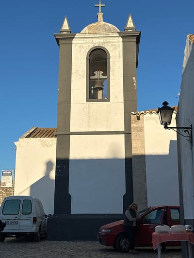 The image shows the bell tower of the Igreja Matriz de Vila Real de Santo António in Vila Real de Santo António, Portugal. The tower is white with dark gray trim and a dome topped with a cross. A bell is visible inside the tower. The tower is situated next to a white building and a street, with a red car and a white van parked on the right side of the image. A woman is near the red car. A black lamppost is attached to the white building. The sky is clear and blue.