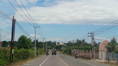 This is a medium shot of a two-lane asphalt road in Lhokdagang, Indonesia. The road is bordered by lush green vegetation and trees on both sides. Several power lines stretch across the frame, supported by tall utility poles. A mosque with a large dome and two minarets is visible in the distance, nestled among the trees.

Vehicles are present on the road. A truck is in the center lane, moving away from the camera. To the left, two motorcycles are also traveling in the same direction, with riders on each. To the right, another motorcycle is approaching the camera, with a rider.

On the right side of the road, a pink building with a sloped roof is partially visible, behind a fence. Further down the road, another building with a green dome-like structure and a water tank on top is seen. The sky is blue with scattered white clouds. The overall scene depicts a rural or suburban setting with evidence of both infrastructure and religious architecture.