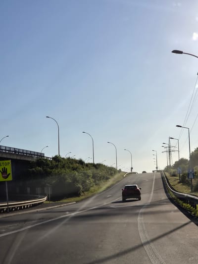 A road is visible in Dudelange, Luxembourg. A red car is on the road with tire marks behind it. Streetlights line the road. The sky is light blue. Green shrubbery is alongside the road. A pedestrian bridge is visible with a yellow sign with a hand and the word "STOP" in block letters. In the distance are electrical towers.