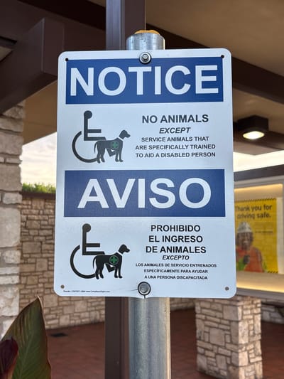 The image shows a bilingual (English and Spanish) sign posted on a metal pole in an outdoor setting, likely a commercial or public area. The sign is white with a blue header that says "NOTICE". Below that, in black text, it states "NO ANIMALS EXCEPT SERVICE ANIMALS THAT ARE SPECIFICALLY TRAINED TO AID A DISABLED PERSON." The sign also features the international symbol of access with a silhouette of a service dog. The Spanish section reads "AVISO" in a blue header, followed by "PROHIBIDO EL INGRESO DE ANIMALES EXCEPTO LOS ANIMALES DE SERVICIO ENTRENADOS ESPECÍFICAMENTE PARA AYUDAR A UNA PERSONA DISCAPACITADA." The background shows a building with stone and dark brown trim, and a glimpse of a yellow sign with text that reads "Thank you for driving safe." The overall scene suggests a location where service animals are permitted, but other animals are not. The specific location is a parking lot in Sonora, United States.