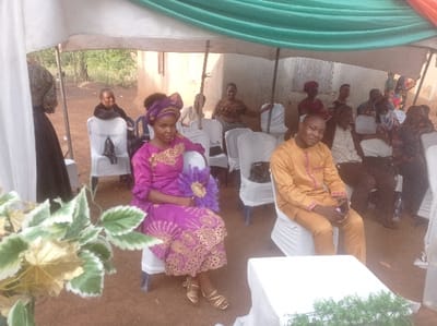 This image depicts an outdoor gathering in Ugbongha, Nigeria, under a temporary tent structure with white and green fabric overhead. The ground is reddish-brown dirt. In the foreground, a woman, appearing in her 20s or 30s, is seated on a white-covered chair to the left of center. She wears a magenta-purple traditional outfit with elaborate gold embroidery on the chest and skirt, a matching headwrap, and gold sandals. She holds a purple decorative hand fan in her lap and looks directly at the viewer. To her right, a man, appearing in his 30s or 40s, sits on another white-covered chair. He wears a golden-yellow traditional tunic and trousers with light embroidery and looks towards the camera with a slight smile.
Behind these two individuals, multiple rows of other attendees are visible, seated on identical white-covered chairs. Their attire varies, including traditional wear and casual clothing. A short, white-draped table with a green leafy decoration occupies the lower right foreground. In the far left foreground, a white and green leafy plant arrangement is partially visible. The background reveals a light-colored building with an open doorway and sparse vegetation, indicating a rural or semi-urban setting. The overall scene suggests a community event or celebration.