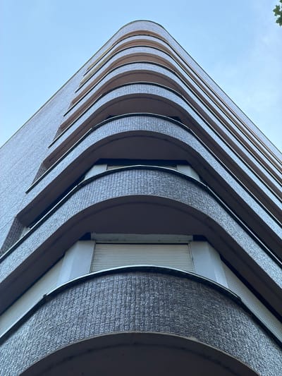 This is a low-angle, close-up shot of a modern building with a curved facade. The building is made of light-colored brick with dark, curved balconies. The balconies have a textured surface that appears to be stone or a similar material. The windows within the balconies are white and have vertical blinds. The shot emphasizes the repeating pattern of the curved balconies and the clean lines of the architecture. The sky is a clear, pale blue, suggesting it is daytime, likely late morning or early afternoon, with fair weather. The upward angle gives a sense of scale and the imposing nature of the structure. There is no text visible in the image. The scene is urban, characterized by modern architecture. The focus is on the abstract architectural forms rather than any activity or people.