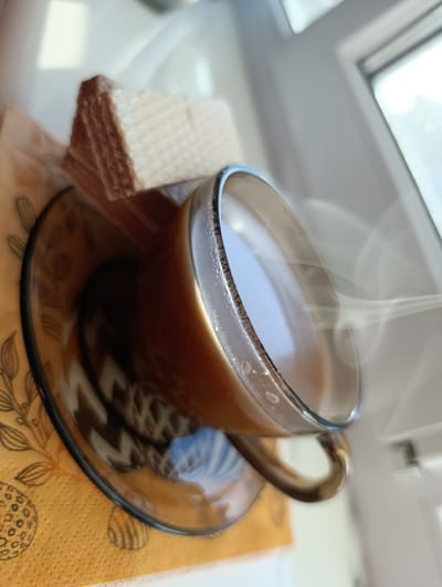 A hot beverage in a clear glass mug sits on a saucer, which is placed on an orange patterned napkin. A stack of wafers rests on the napkin next to the mug. Steam rises from the drink, indicating it is hot. The scene is set on a windowsill, with natural light illuminating the objects. The background shows a window frame and the bright outdoors, suggesting an indoor setting with a view. The image is taken from a close-up, slightly tilted perspective. There are no people present in the image. The location is identified as Pănășești, Moldova.