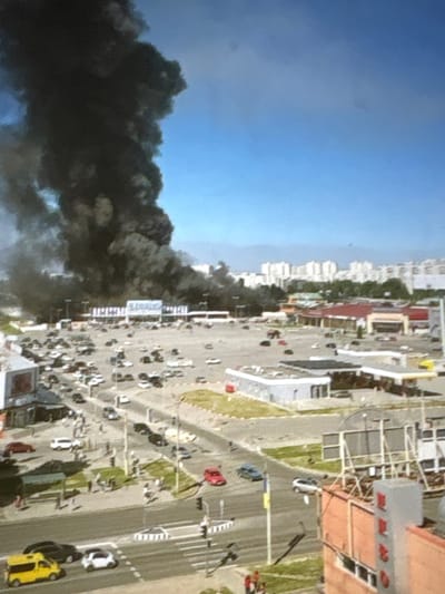 This aerial view captures a chaotic scene in Pori, Finland, marked by a large plume of black smoke billowing into a clear blue sky. The smoke originates from a densely populated urban area, suggesting a significant fire or explosion. Below the smoke, a bustling street is visible, lined with buildings of varying heights, including apartment complexes and commercial structures.
Numerous vehicles, including cars and what appear to be trucks, are scattered across the scene, some parked and others in motion. Pedestrians can be seen, though their numbers are difficult to ascertain precisely due to the image's resolution and distance. The overall impression is one of urgency and potential danger, with the dark smoke contrasting sharply against the bright daylight and blue sky, indicating the event is occurring during the day.
The architectural style of the buildings suggests a European setting, consistent with the provided location of Pori, Finland. There are some visible signs and text on the buildings, but they are not clear enough to be read. The presence of traffic and people in the vicinity of the smoke suggests the incident may have disrupted daily life and could be a cause for concern among the local population. The scene appears to be set in a developed urban environment with a mix of residential and commercial properties.