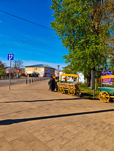 The image depicts a bright, sunny day in Uglich, Russia. The foreground is dominated by a paved sidewalk made of rectangular blocks. Two wooden carts with large yellow wheels are positioned on the right side of the sidewalk, near a tree with green leaves. The first cart appears to be a vendor stand with items displayed on top, while the second cart has a green covering.
A person in a dark coat is standing near the carts. Across the street, there is a row of buildings, primarily in yellow and light colors. A signpost with a blue sign indicating two-way traffic is visible on the left side of the image. A string of decorative lights is suspended above the road. A metal fence runs along the roadside. The sky is a clear, deep blue.