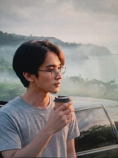 A medium shot captures a young Asian man with dark hair and glasses, holding a coffee cup to his lips, in what appears to be a scenic overlook in Cigadung, Indonesia. He is wearing a gray t-shirt and is positioned to the left of the frame. Behind him, a blurred landscape of rolling hills shrouded in mist stretches towards a hazy sky. The man is partially leaning against a car, the side of which is visible on the right side of the image. The overall lighting suggests it might be dawn or dusk, with a soft glow illuminating the scene.