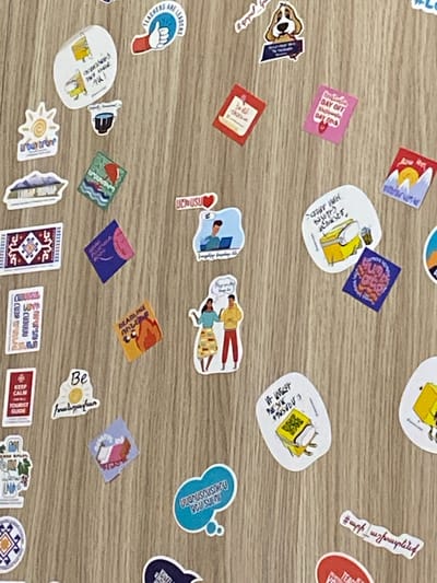 This media file displays a collection of stickers scattered on a wooden surface. The stickers feature a variety of subjects, including illustrations of people, objects, and abstract designs. Some stickers depict cartoon characters, while others have text in what appears to be Armenian and English. There are also stickers with images of brains, computers, luggage, a sun, and various patterned designs. The overall scene suggests a casual or creative environment, possibly a desk or table where these stickers have been placed. The wooden surface provides a neutral background, allowing the colorful and diverse stickers to stand out. The presence of stickers like "DAY OFF" and "#teacherlife" hint at a theme related to work, leisure, or personal expression. Some stickers have more detailed illustrations, such as a scene with people working on computers, or a depiction of a woman with a sun motif. The arrangement of the stickers is somewhat random, with no clear organization. The lighting suggests an indoor setting, likely during the daytime.