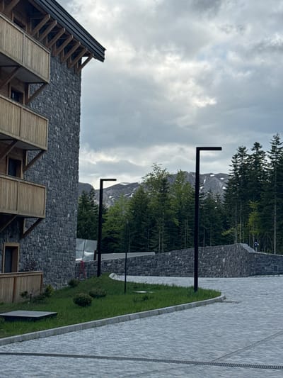 The image shows a mountainside landscape in Katun Bačko brdo, Montenegro, on an overcast day. A modern stone building with wooden accents is in the foreground. Tall, thin evergreen trees, along with a rocky mountain peak with snow, are behind the building. Several black, L-shaped lampposts are between the building and trees. A paved path with a strip of green grass is alongside a stone wall. The sky is covered in gray clouds.