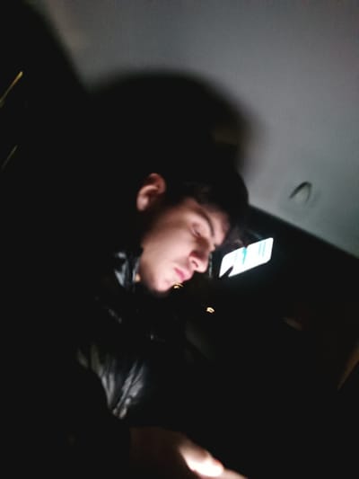 The image depicts a low-light, candid portrait of a young person, likely male, situated within what appears to be the interior of a vehicle, such as a car or van, in Uckange, France. The subject, visible from the chest up, has dark hair and is positioned facing downwards and slightly to the right, with their profile illuminated.
The primary light source originates from a screen held in the subject's hands, casting a soft, warm glow onto their face, particularly highlighting their chin, lips, and nose. Their eyes are downcast or closed, suggesting focused attention on the device, relaxation, or perhaps drowsiness. They are wearing a dark, possibly black, jacket with visible texture, which could be leather or a synthetic material.
The background is predominantly dark, indicating that the scene takes place at night. The ceiling of the vehicle is visible in the upper part of the frame, appearing light grey. In the upper right quadrant, a reflective surface, possibly a rear-view mirror or a sun visor, reflects a bright, rectangular light source that shows vertical lines, suggestive of a digital display, though no specific text is discernible. Other blurry, faint lights, possibly external or internal reflections, are scattered in the dark background. The overall impression is one of quiet engagement with a mobile device during nighttime travel or waiting.