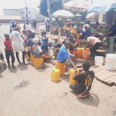 The scene is a street market in Kufang, Nigeria, located at latitude 9.863 and longitude 8.867. There are many people, mostly children, sitting on yellow plastic containers. Some children are playing with the containers, while others are just sitting and watching. There is a wooden platform with various items, possibly fruit, on display. A woman in a striped shirt is standing in front of a stall with a selection of fruit. There are many people in the background of the image, who appear to be either shopping or watching the children.