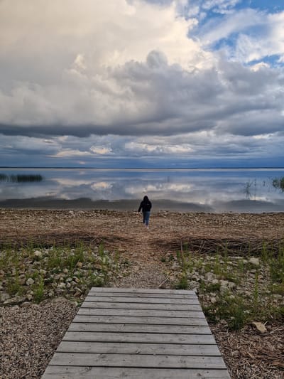 A person wearing a dark hoodie and light-colored pants walks away from the viewer towards a calm body of water. They are walking on a wooden boardwalk that transitions into a gravelly shore. The water's surface is highly reflective, mirroring the dramatic, cloudy sky above. Tall reeds or grass are visible along the water's edge and in the distance. The scene appears to be a natural environment, possibly a lake or large pond, on the outskirts of Īdeņa, Latvia. The overall mood is serene and contemplative, with the vast sky and reflective water dominating the composition.