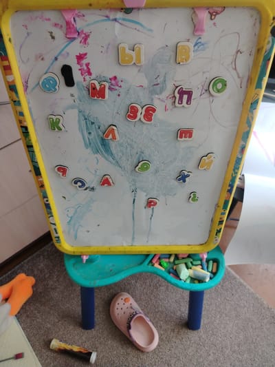 The image shows a child's magnetic whiteboard with various magnetic letters and drawings on it, positioned on a stand. A tray attached to the stand holds an assortment of colorful chalk. Scattered around the base of the stand are a pink shoe, a tube of effervescent tablets, and an orange object that appears to be a child's toy. The scene suggests a home environment where a child has been engaged in drawing and play. The location is specified as Тулаўскі сельскі Савет, Belarus, implying a rural or semi-rural setting within the country.