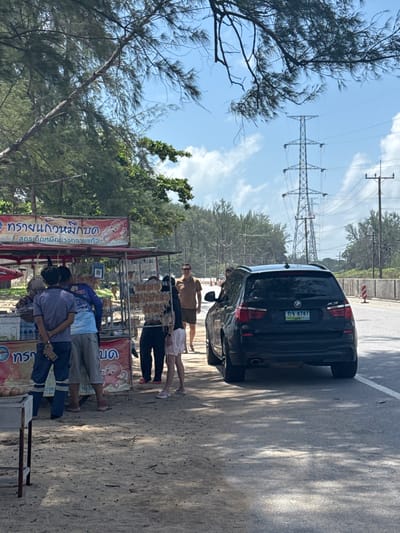 A black BMW X3 SUV is parked on the side of a road in Ban Tha Chathai, Thailand. The vehicle is facing away from the camera, and its license plate reads "กจ 8787". To the left of the vehicle, a food cart is set up on the sandy ground, displaying what appears to be dried squid or other seafood. Several people are gathered around the cart, some interacting with the vendor. Behind the food cart, a line of trees borders the roadside. In the background, tall electricity pylons and power lines are visible against a bright, partly cloudy sky. The scene appears to be a roadside market on a sunny day.