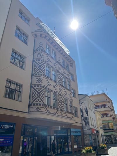 The image is a ground-level, eye-level, wide-angle outdoor photograph taken on a sunny day, looking up at the facades of several buildings on a city street. The architecture features a mix of styles, with the prominent building on the left displaying a distinctive art deco or modernist design with geometric patterns in brown and cream. This building has multiple floors with rectangular windows and a curved section with a glass roof canopy. Above the first floor, there is a sign that reads "ELEKTRONICKÉ CIGARETY". Further down the street, other buildings are visible with various shopfronts at the ground level. One shop has a sign that says "LIQUA", and another displays "PAPÍRNICTVÍ". A large black and white mural of a person wearing a beret is visible on the facade of a building in the middle ground. The sky is bright blue with a strong sun creating lens flare. There are some potted plants with yellow flowers in concrete planters on the sidewalk in the foreground. A few people are present on the street, including one individual walking away from the camera on the sidewalk in front of the shops. The image appears to have been taken in Náchod, Czechia, based on the signage.