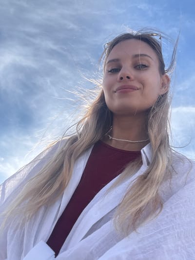A photograph taken from below captures a young woman with long, blonde hair. She is wearing a burgundy top with a white overshirt. Her face is visible, and she has a delicate necklace around her neck. The background is a blue sky with scattered clouds. The location cannot be determined from the image content alone.