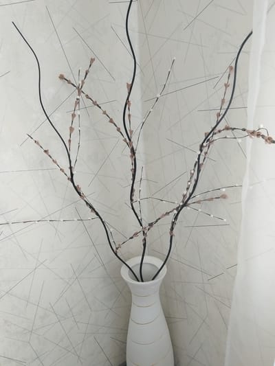 A white, flared vase with gold horizontal bands holds a decorative arrangement of thin, dark branches with small, light pink bud-like accents and white spherical embellishments. The branches are sparse and extend upwards and outwards. Behind the vase and branches is a wall with a textured, light-colored wallpaper featuring a pattern of thin, dark, intersecting lines of varying lengths. To the right of the arrangement, a sheer white curtain is visible, draped and slightly opaque. The scene appears to be an indoor setting, possibly a home or commercial space, in the city of Andzhero-Sudzhensk, Russia. There are no people or animals present in the image.
