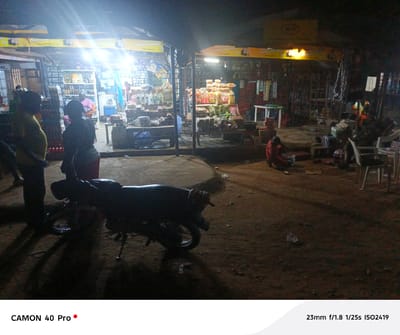 The photograph captures a nighttime scene in Langtang, Nigeria, depicting a gathering around two small commercial storefronts. These shops are brightly illuminated, revealing displays of various goods. Banners advertising the brand "CHOOSE MALTINA CHOOSE HAPPINESS" are prominently displayed above the shops.
In the foreground, to the left, figures stand near a parked motorcycle, their forms largely silhouetted against the light. To the right, individuals are seated on chairs and on the ground, engaged in possible social interactions. The ground appears uneven and unpaved. The overall impression is that of a community gathering spot after dark in a developing urban setting.
