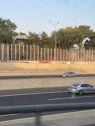 This image depicts a section of a multi-lane highway in Kefar Sava, Israel, under a clear, bright sky. The foreground shows a dark, blurred horizontal element, possibly the interior of a vehicle or a barrier. The highway features at least three visible lanes of asphalt, separated by white double solid lines, with a single yellow line on the far left. Concrete barriers define the highway's edges and separate different levels of traffic flow.

Above the main highway lanes, a higher concrete retaining wall is visible, adorned with graffiti that includes a pink, stylized word resembling "Sloam" and other indistinct lettering such as "ZNA." Behind this wall, a sound barrier composed of vertical, light-colored posts with transparent or translucent panels stretches across the scene.

Beyond the sound barrier, dense green trees and foliage are present, partially obscuring residential or commercial buildings with flat roofs. A tall, light-grey street light pole with two luminaires extends vertically from behind the sound barrier into the sky on the right side of the image.

Two silver/grey sedans are visible on the highway, both appearing to move from right to left across the frame. One vehicle is in the lower right lane, and another is on a higher, more distant lane. No pedestrians or other human activity are observed. The overall environment suggests an urban or suburban setting with significant infrastructure.