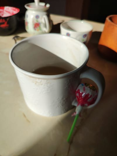 A close-up shot features a white ceramic mug filled with amber liquid, likely tea, positioned on a light-colored, speckled countertop. A small, decorative lollipop with red and white wrappers and a green stick is placed in the mug's handle. In the background, several other ceramic containers are visible, one with a floral pattern and a lid, and another smaller bowl also decorated with blue and red flowers. The lighting suggests it is daytime, with sunlight casting shadows across the scene. The overall impression is of a casual, home setting, possibly a kitchen or dining area, located in Буйніцкі сельскі Савет, Belarus.