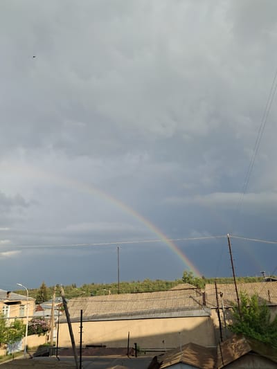 The image captures a view of a residential area in Gavar, Armenia, under a cloudy sky that features a prominent rainbow. The foreground shows rooftops of several buildings, with corrugated metal and tile materials visible. Electrical poles and wires crisscross the scene. In the mid-ground, a line of trees and hills forms the backdrop, with some houses and structures nestled amongst them. A solitary bird is visible in the upper left portion of the sky. The overall atmosphere suggests a recent or ongoing rain shower, common in Gavar's climate.