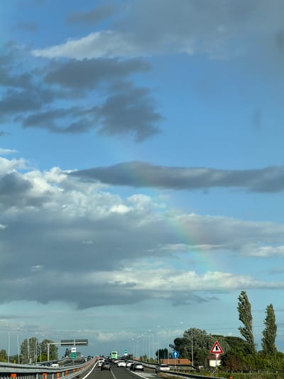 The image shows a view over a section of highway in Vecchiano, Italy. The sky is predominantly blue with various cloud formations including a faint rainbow segment. Alongside the highway is a metal guardrail and trees and shrubbery. Road signs are visible, along with a line of vehicles on the highway. Street lights line the road.