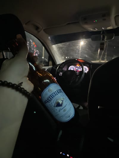 A first-person perspective shot shows a person's hand holding a bottle of beer inside a car at night. The person is wearing a black beaded bracelet on their wrist. The car's dashboard is visible, with illuminated gauges and lights. The bottle has a label with Cyrillic text and the word "Premium" printed on it. The exterior of the car is dark, with streetlights reflecting on the windshield. The scene suggests the person is a passenger or driver in a vehicle at night in Kizner, Russia. The lighting indicates it is nighttime, and the presence of streetlights implies an urban or suburban setting. There are no strong emotional cues, but the act of holding a beer in a car at night could suggest relaxation or socializing.