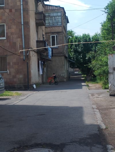 A person rides a bicycle down a narrow street in front of an apartment building in Nor Hachn, Armenia. The building is made of red brick and has balconies and clothes hanging from lines. Trees and a fence line the street on the right side of the image. The sky is blue with some clouds.