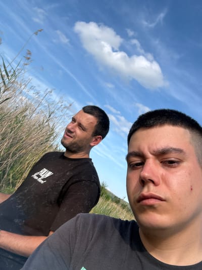 Two males are depicted outdoors in what is identified as Zamfir, Bulgaria, captured in a selfie-style photograph. The subject in the immediate foreground, a young adult male, has short dark hair styled in a buzz cut and a subtle facial hair growth on his upper lip and chin. His expression is neutral to slightly stern, and a small red blemish is visible on his right cheek. He wears a dark-colored short-sleeved t-shirt. Positioned to his left and slightly behind, the second male, an adult, has short dark hair and a beard. He is observed looking towards the left side of the frame with a slight upward curve to his mouth. He wears a black short-sleeved t-shirt featuring white text that reads "FOLLOW Your Heart." The background consists of tall, dried, light brown grasses or reeds and a vibrant blue sky containing scattered white clouds, some appearing wispy and others more voluminous. The lighting suggests a bright, sunny daytime environment.
