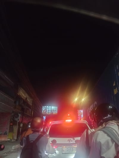 The image shows a street scene at night, likely in Calukan RT 1 RW 5 Keboansikep, Indonesia, given the provided context. The scene is dominated by traffic, with several vehicles visible, including a white car in the foreground and motorcycles. Two individuals on a motorcycle are the most prominent subjects, facing away from the camera. The person in the back is wearing a dark helmet and a light-colored jacket over a backpack, while the person in front has on a lighter brown helmet and a brown jacket. Red taillights of the vehicles create a strong visual element, along with bright streetlights and illuminated signs. The buildings on either side of the street appear to be commercial establishments, with various signs and advertisements. The overall atmosphere suggests a busy urban environment at night, possibly experiencing a traffic jam.
