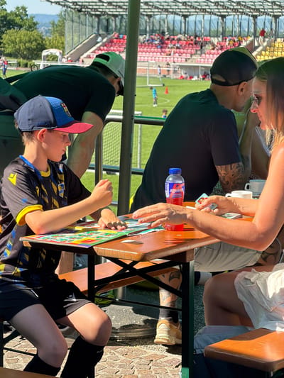 A family is sitting at a table outdoors at a stadium in Fentange, Luxembourg, with a soccer game taking place in the background. A boy wearing a soccer uniform and a Barcelona cap is playing a game on a board. A woman with long blonde hair and a man with a black baseball cap and arm tattoos are sitting across from the boy, playing cards. A water bottle is on the table. A person with a white baseball cap is sitting behind the boy. Red stadium seating and a soccer field are visible in the background.