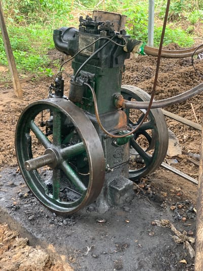The image shows a large, green, industrial engine, lying on its side, partially obscured by green foliage in the foreground. The engine has a large, spoked, black wheel. The engine is lying on a bed of dark, wet, muddy soil. The engine appears to be in disrepair, with parts missing or damaged. The image was taken at coordinates (latitude: 7.119, longitude: 5.362) in the city of Egbeda, Nigeria.