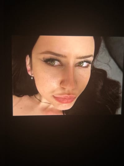 The image is a close-up, low-angle shot of a woman's face. She has dark hair, fair skin with freckles, and green eyes. She is wearing dark eyeliner and mascara, and has light pink lipstick on. A small, silver earring is visible in her left ear. The background is dark and out of focus, with a hint of gray fabric on the right side. There is no visible indication of the city of Korostyshiv, Ukraine in the image.