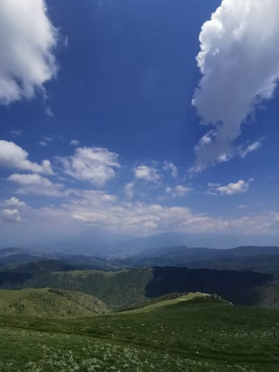 A high-angle, wide shot captures a panoramic view of rolling green hills under a vast blue sky dotted with white clouds. The foreground features a lush, grassy slope with sparse, light-colored vegetation. In the mid-ground, the terrain transitions into more densely forested hills, with a noticeable shadow cast across them from a cloud. Beyond these, a hazy expanse of distant mountains and a valley with a faint hint of structures suggest a settlement or town. The sky is a dominant feature, with large, fluffy clouds dominating the upper half of the frame, contrasting with the deep blue of the sky. The overall impression is one of natural beauty and expansive scenery. This scene is located in the vicinity of Brestovo, Bulgaria. There are no discernible people or actions depicted in the image.