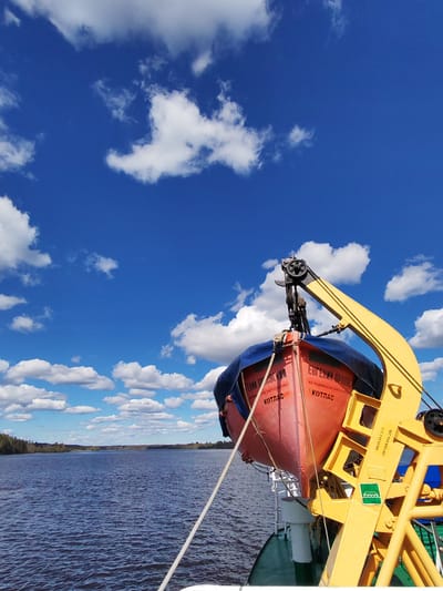 The image shows a lifeboat hanging on a crane on a ship, with a body of water and trees visible in the background. The sky is blue with scattered white clouds. The lifeboat is orange and has some Russian text on its side including "Kotlas", referring to the city in Russia. The crane is yellow and black. The water appears calm.